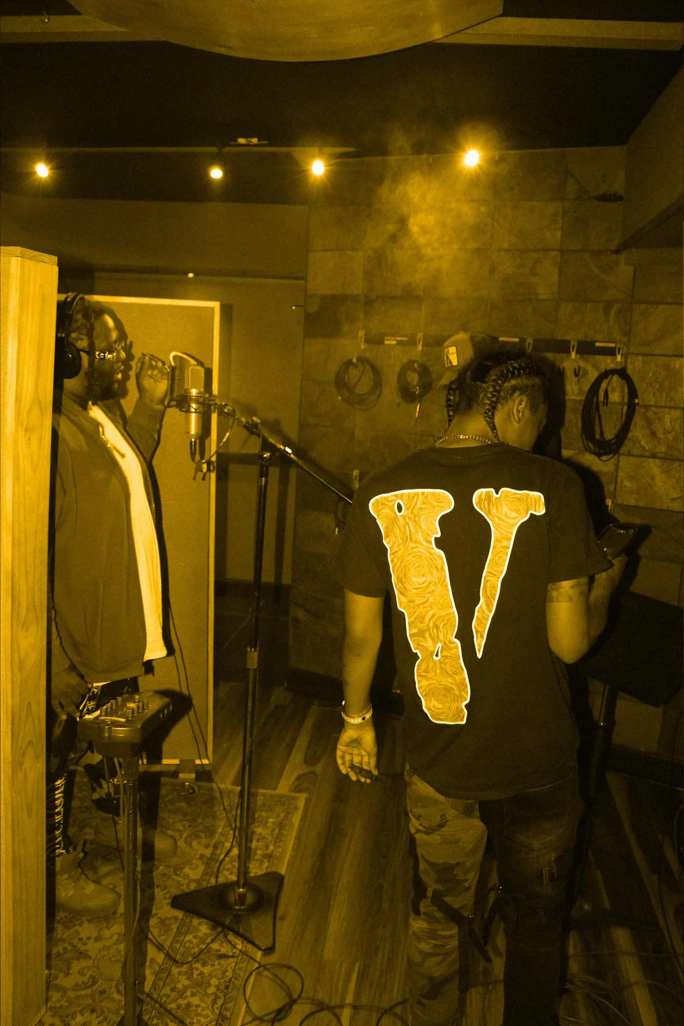 Two artists in a smoky vocal booth, one recording, one reading lyrics